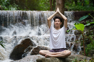 Man meditating and practicing yoga on rock in forest near waterfall. Sport, yoga, pilates, fitness, healthy lifestyle concept.