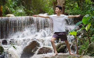 Attractive young man practicing yoga meditation and breathwork   in forest near waterfall. Yoga, pilates, healthy lifestyle concept.
