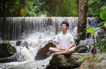Fototapeta na wymiar Peaceful young man practicing yoga meditation near waterfall in forest. Concept of calm and meditation.