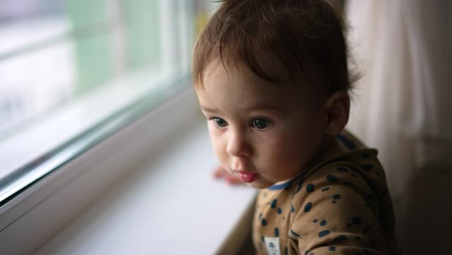 Cute baby standing at the window and banging by the glass. Lovely kid turning around and looks at camera. Close up.