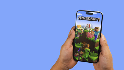 Fototapeta premium Child holding a smartphone iPhone 14 Pro with Minecraft mobile game app on the screen. Blue background. Rio de Janeiro, RJ, Brazil. September 2022