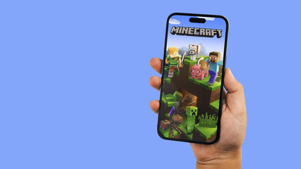 Fototapeta premium Child holding a smartphone iPhone 14 Pro with Minecraft mobile game app on the screen. Blue background. Rio de Janeiro, RJ, Brazil. September 2022