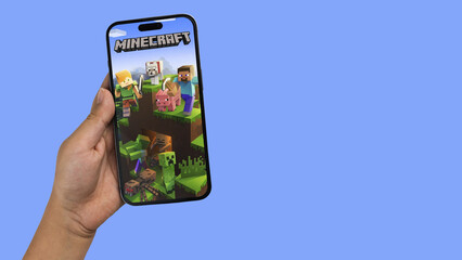 Fototapeta premium Child holding a smartphone iPhone 14 Pro with Minecraft mobile game app on the screen. Blue background. Rio de Janeiro, RJ, Brazil. September 2022