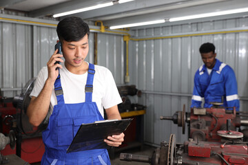 Multiethnic technician mechanic using metal lathe machine operate polishing car disc brake with smart mobile phone and clipboard at garage. Maintenance automotive and inspecting vehicle part concept