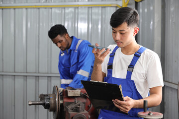 Multiethnic technician mechanic using metal lathe machine operate polishing car disc brake with smart mobile phone and clipboard at garage. Maintenance automotive and inspecting vehicle part concept