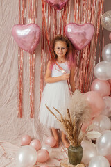 Little girl in a white dress poses on a festive pink background with sparkles and heart-shaped balloons. Valentine's Day celebration. Happy birthday. 