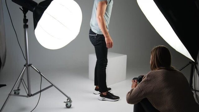 Male model posing for a photographer in studio. Woman taking pictures of man's footwear on white backdrop.
