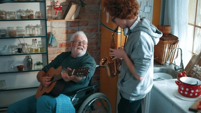 Cheerful old person with a disability in a wheelchair plays the guitar together with his grandson who plays the saxophone