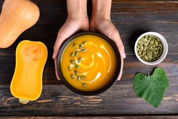 Butternut squash pumpkin soup in bowl holding by hand on wooden background, Homemade food in autumn...