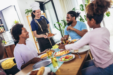 A multi-generational African-American family enjoying food at their dinner table. Caucasian woman cook serve them.