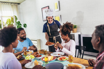 A multi-generational African-American family enjoying food at their dinner table. Caucasian woman cook serve them.