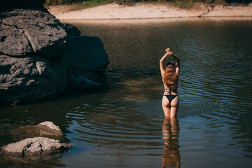 Fototapeta premium A young girl with long blond hair, a slender figure and a beautiful back, in a black swimsuit, stands in a river against the backdrop of rocks.