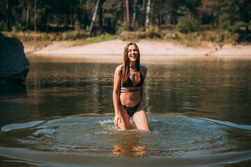 A young girl with long blond hair, a slender figure, in a black swimsuit, enters the water and smiles, against the background of the beach and stones in the river, on a hot summer day.