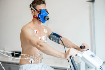 Man athlete with breath mask and electrodes training on bike simulator, examining his cardiovascular system at medical office - obrazy, fototapety, plakaty