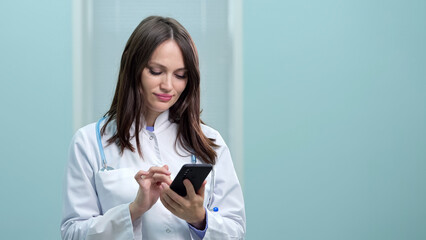 Joyful woman in white lab coat rests during lunch break in hospital office. Brunette doctor chats with friends on social networks holding smartphone in hand
