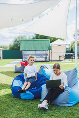 Naklejka premium Mom is on maternity leave. Charming young woman with little son working outdoors in summer park with laptop while sitting on chair on green grass