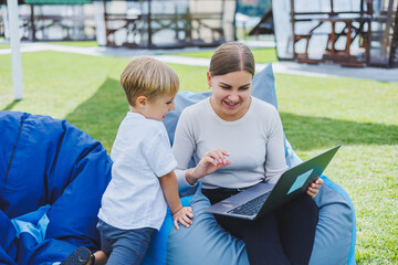 Fototapeta na wymiar Mom is on maternity leave. Charming young woman with little son working outdoors in summer park with laptop while sitting on chair on green grass