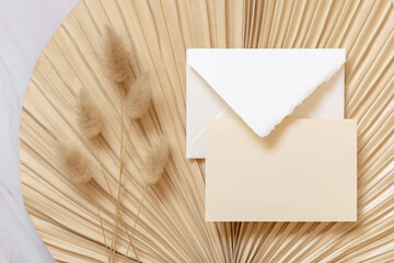 Blank card and envelope on dry beige palm leaf near hare's tail grass top view,  boho wedding mockup