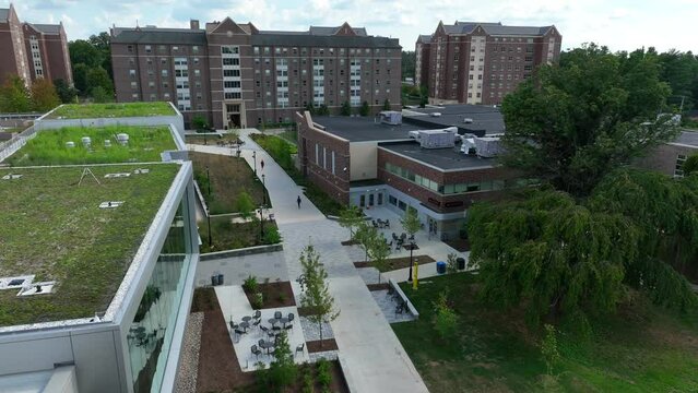 Aerial tilt up shot reveals college dorm building. Academic buildings feature green spaces on roof. Sidewalks make campus walkable for students. Residential university life theme.