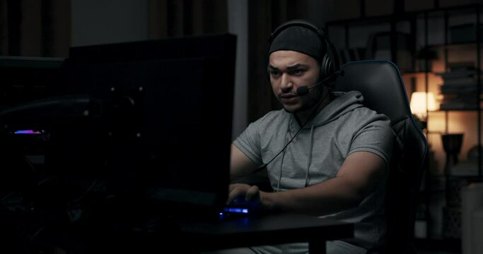 Man with beard plays online game on computer. Guy is wearing cap and headphones. He is wearing gray T-shirt. Player sits in dark room, communicates with opponents and presses on keyboard.