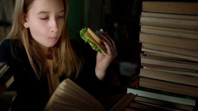 Teenager girl sits in library, reads book and eats sandwich. Student receives new knowledge. Smart and well-read person. Education and development. Interesting school literature. Leisure activity.