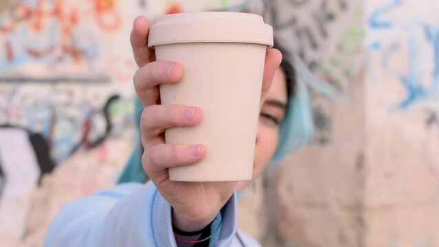 Teenager girl in blue oversize hoodie showing coffee to go cup. Blue haired teen girl staying outdoors near graffiti wall. Mockup, head shot. Adolescence concept