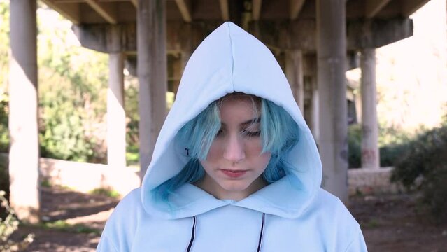 Teenager wearing blue oversize hoodie looking down. Blue haired teen girl staying outdoors against bridge pillars. Head shot. Hipster and adolescence concept