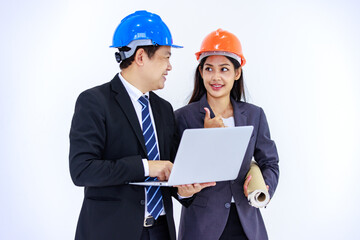 Millennial Asian successful professional male and female engineer in formal suit and safety hard helmet standing together holding blueprint looking at laptop notebook computer on white background