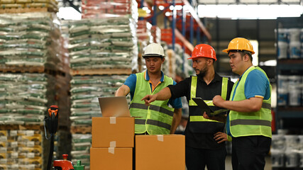 Male supervisor and warehouse workers in hardhats and reflective jackets checking stock and order details on laptop computer