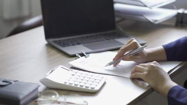 Young Asian businesswoman presses calculator to calculate income tax and corporate income in private office. Finance, accounting, marketer's statistical graph.