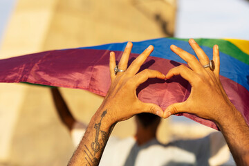 Hands making heart sign in front of rainbow flag.