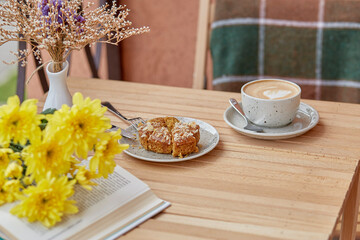 Aesthetic autumn lunch - coffee time for yourself book and dessert. Cutting doughnut with cappuccino among chrysanthemum. Atmospheric lunch, guilty pleasure.