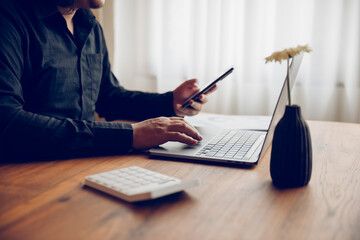 A young man working using a smartphone and a laptop computer. Man's hand using smartphone inside white room building