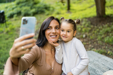 Pregnant white woman and little girl taking selfie together while sitting on bench in park. enjoy beautiful fall day.