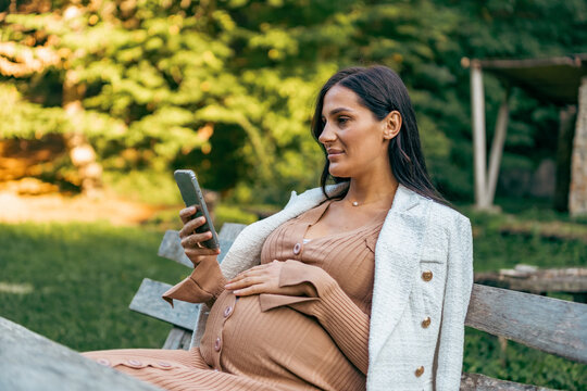 Pregnant woman sits on bench in the park and using phone. Happy female have rest outdoor during the walks