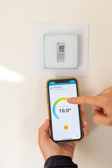 Person holding a smartphone in their hands to adjust their connected thermostat in a French house - 534467860