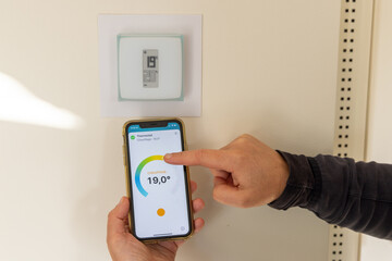 Person holding a smartphone in their hands to adjust their connected thermostat in a French house - 534467869