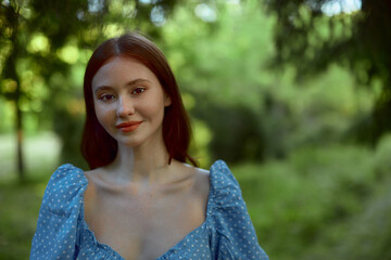 Young pretty woman in blue dress with red hair looking at the camera and smiles. The girl posing and enjoying walk in the woods.