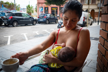 close up of young mom breastfeeding newborn baby daughter in public outdoors in the street at a bar drinking coffee - obrazy, fototapety, plakaty