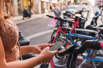 Image of an unrecognizable woman using her phone terminal to unlock an electric bike for rent in the big city of Madrid.