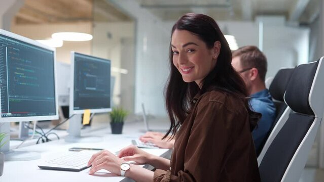 Rear of young pretty woman IT programer working on desktop computers typing on keyboard coding a program turning to camera and smiling. Close up of professional worker in modern tech company