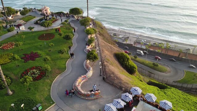 Drone video of a park on cliff edge with the ocean below and a coastal freeway. Drone tilts up and turns left revealing grass, palm trees, people walking around. Recorded in Lima, Peru in Miraflores.