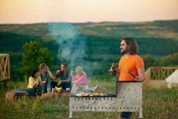 A middle aged man is laying down lots of different types of vegetables onto an outside grill and smiling proudly as he is with his friends in the nature. - Powered by Adobe