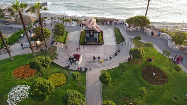 Drone 4k video of "Parque del amor" park in Miraflores, Lima, Peru. Drone flies forward. Below bushes, grass, palm trees and statue of a couple kissing. Below is the beach and ocean. During sunset.