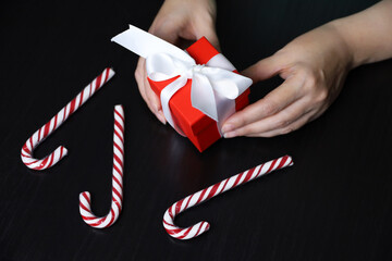 Red gift box in female hands on dark wooden table with candy canes. Concept of Christmas holiday, romantic surprise