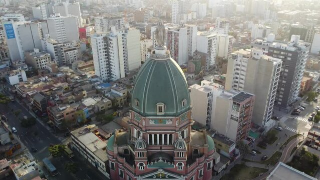 Drone video of a church in Lima, Peru. Drone flies back showing the church and buildings in the background. Recorded around sunset, light shines on the virgin mary statue on top of the church's dome.