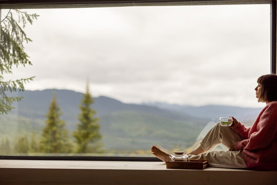 Woman sits with phone on window sill and enjoys scenic view on mountains while resting in wooden house on nature. Recreation and escaping to nature concept