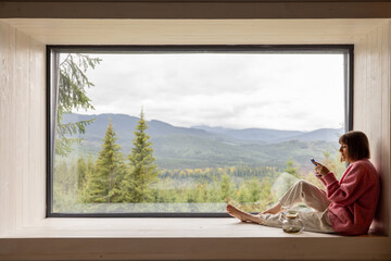 Woman sits with phone on window sill and enjoys scenic view on mountains while resting in wooden...