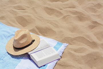 Beach towel with open book and straw hat on sand, space for text