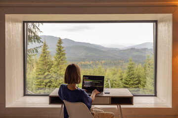 Woman works on laptop while sitting by the table in front of panoramic window with great view on mountains. Remote work and escaping to nature concept - 534496839